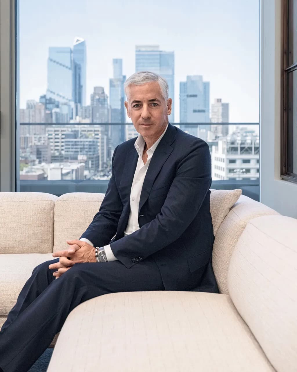 Bill Ackman sitting on a couch with the New York City skyline visible through the window behind him
