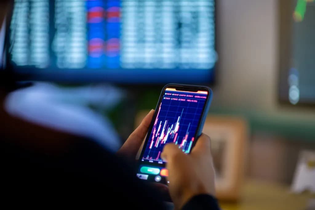 A person checking cryptocurrency price charts on a smartphone with a trading screen blurred in the background